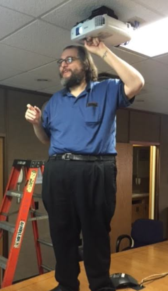 Tom Backus standing on a conference table while installing a ceiling-mounted projector